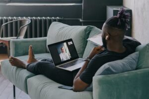 African American man relaxes on a couch while video chatting on a laptop, enjoying a casual day indoors.