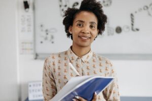 Confident female teacher smiling while holding a notebook in a classroom setting.